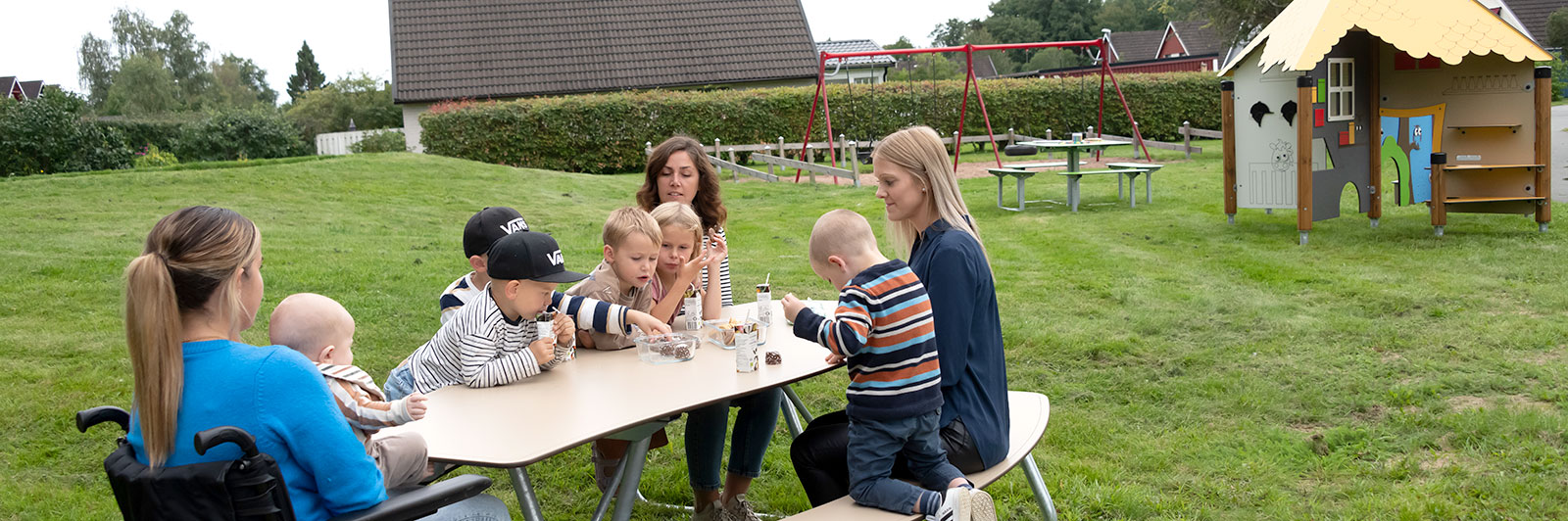 A group of people with children gather round an inclusive picnic table near a playground.